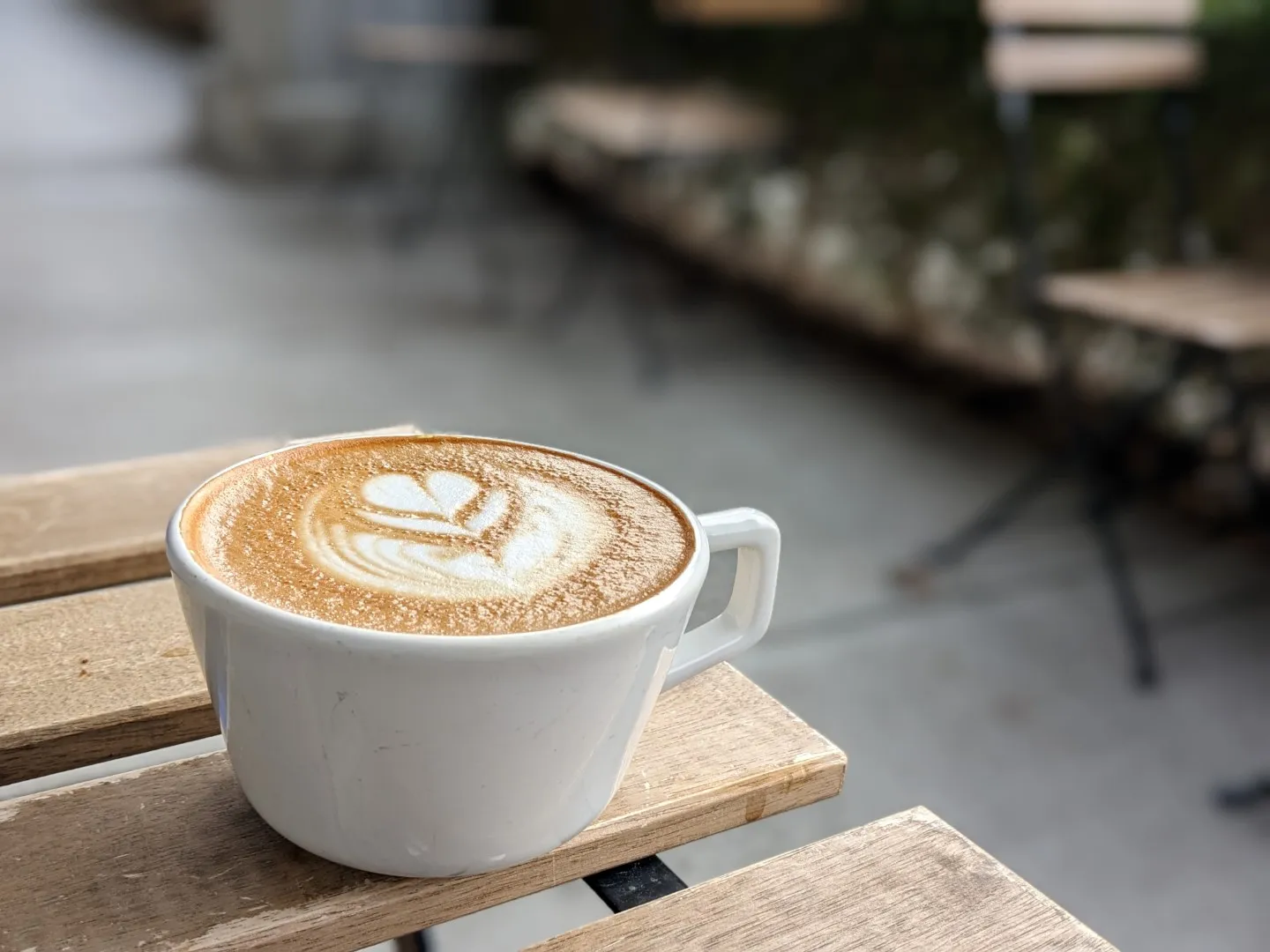 A picture of a beautiful cappuccino with leaf-like latte art sitting on wooden slatted table outside