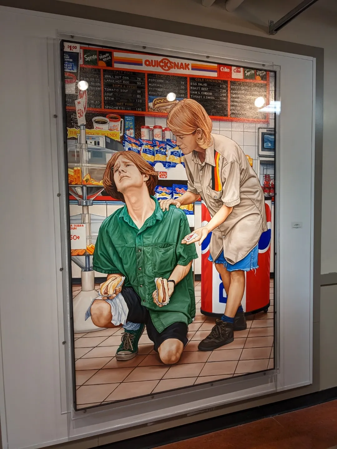 a painting by Chris Woods titled "The Realm of Convenience II". It depicts a blonde man on his knees with hotdogs in his hands being consoled by another blonde woman with ketchup in her hand. They&#x27;re located in a convenience store.